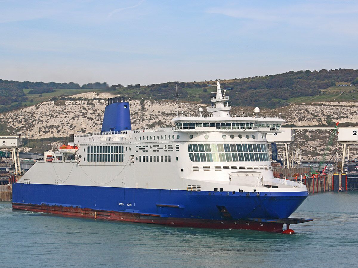 Große blau-weiße Fähre fährt durch einen Hafen mit weißen Klippen und grünen Hügeln im Hintergrund.