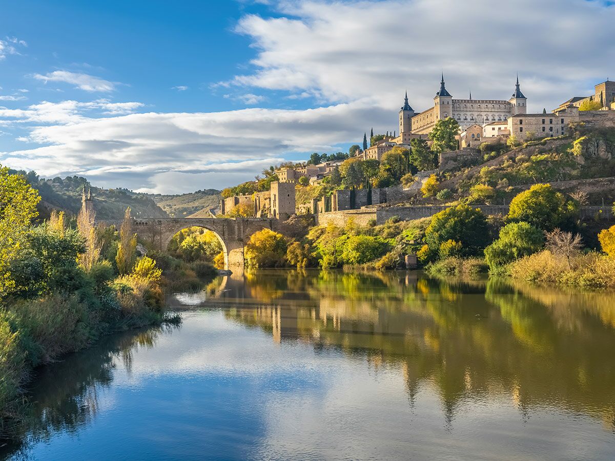 Toledo, Spanien: Historische Stadt mit Alcázar, Fluss Tajo und San Martín Brücke unter blauem Himmel mit Wolken, Herbstlaub.