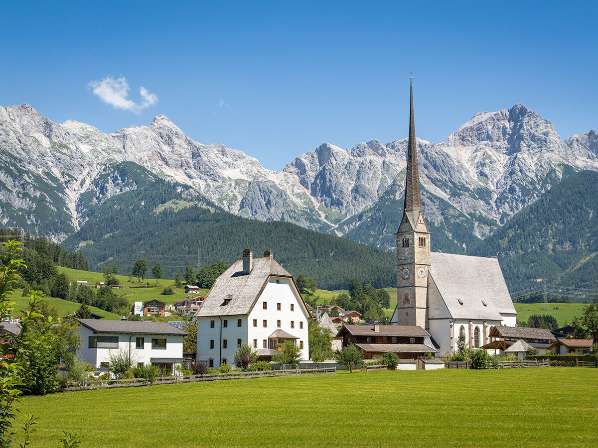 Idyllisches Bergdorf mit hoher Kirche, traditionellen Häusern, grünen Wiesen und majestätischen Alpen unter blauem Himmel.