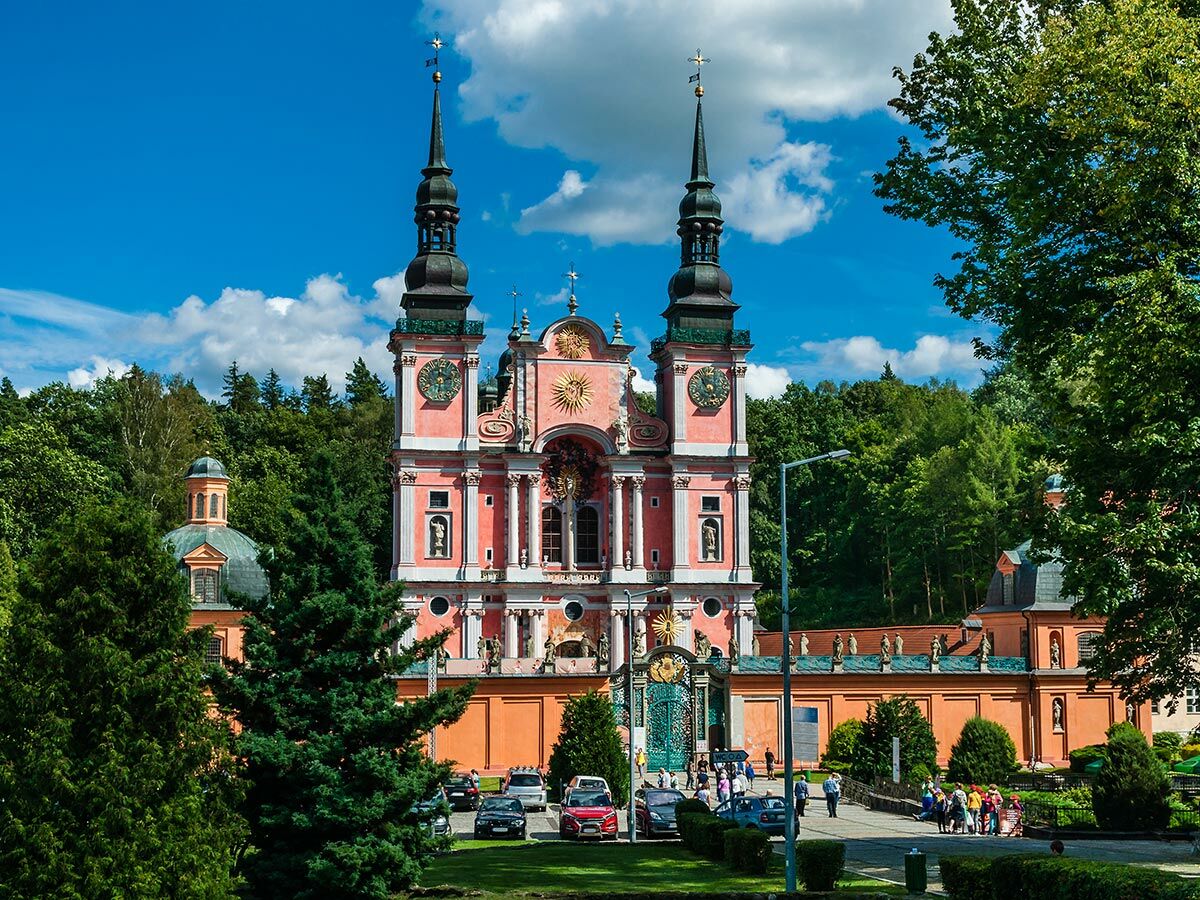 Historische Barockkirche in Krzeszów, Polen, mit rosa Fassade, zwei hohen Türmen, Uhren und Statuen, umgeben von grünen Bäumen.