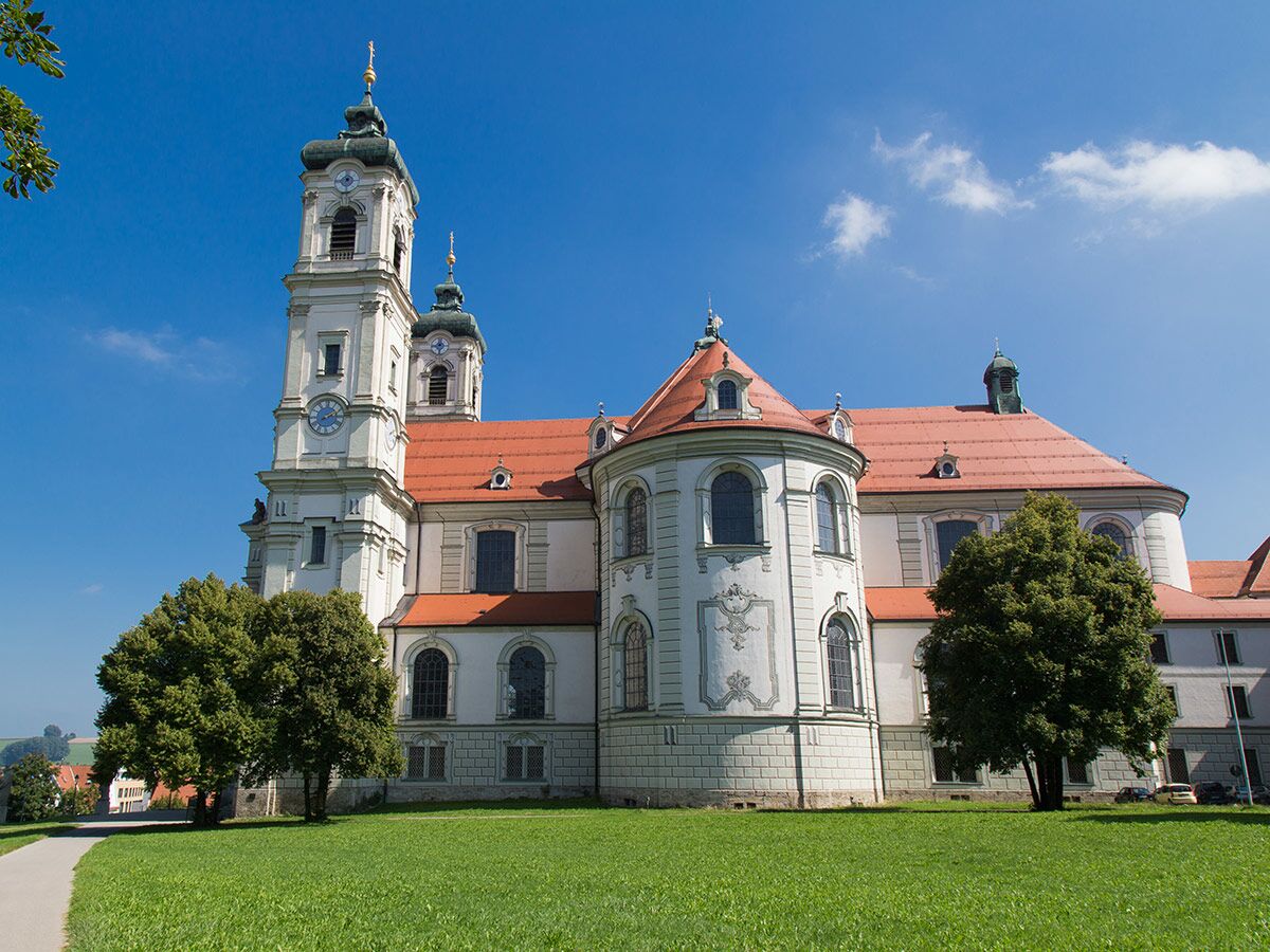 Barockkirche der Abtei Ottobeuren bei Sonnenschein und blauem Himmel, mit grüner Wiese und Bäumen im Vordergrund.