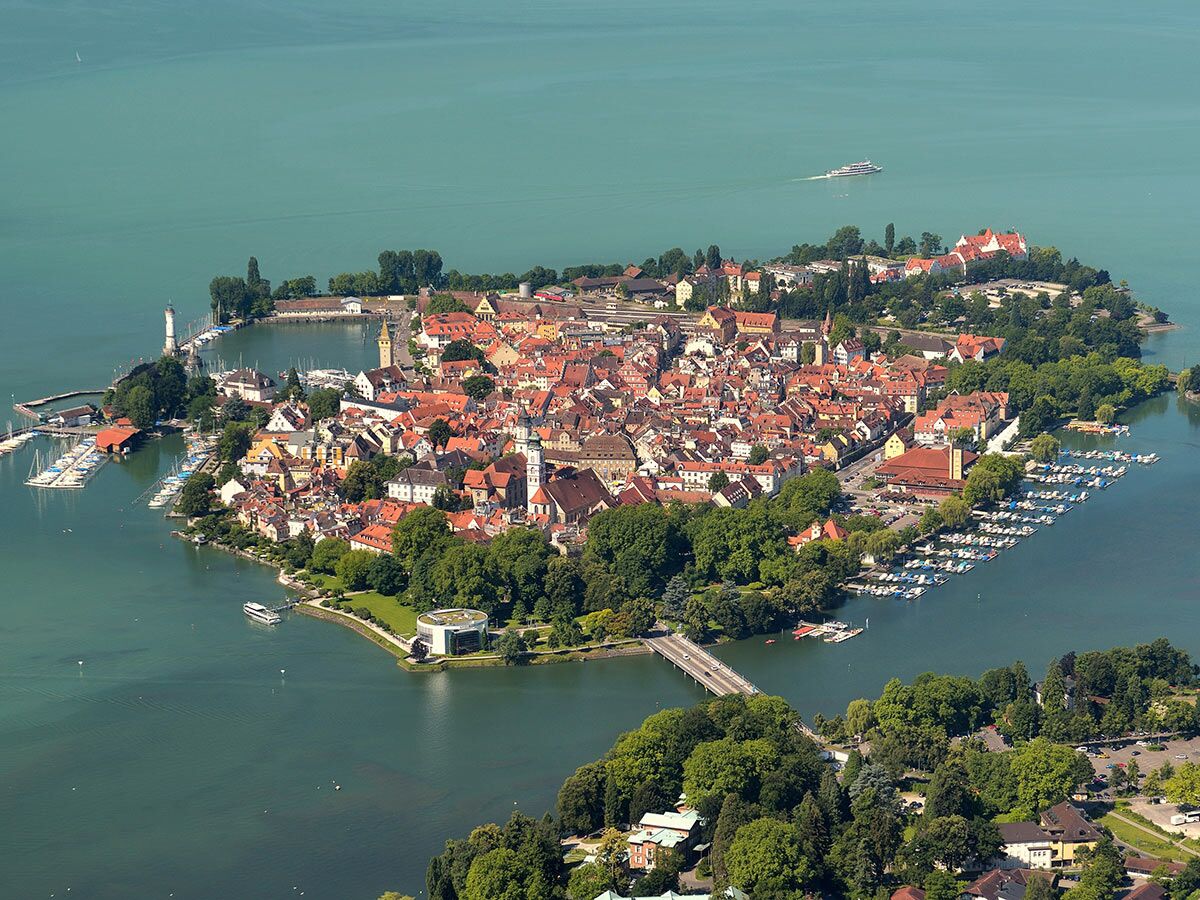 Luftbild der Inselstadt Lindau im Bodensee mit Leuchtturm, Hafen, roten Dächern und grünen Bäumen an einem sonnigen Tag.