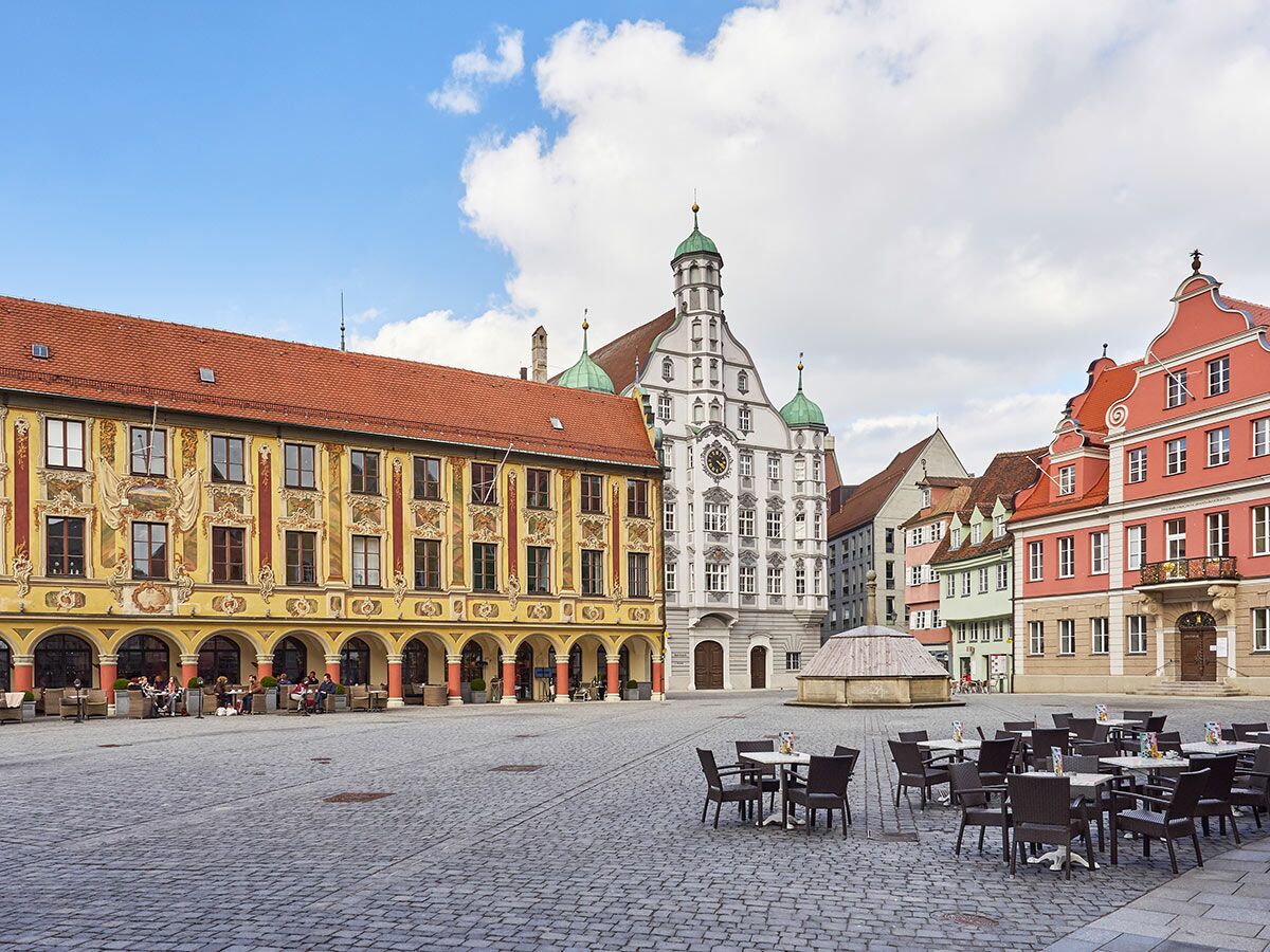 Historischer Marktplatz Landsberg am Lech mit Rathaus, bunten Häusern, Brunnen, Cafés unter Arkaden und blauem Himmel.