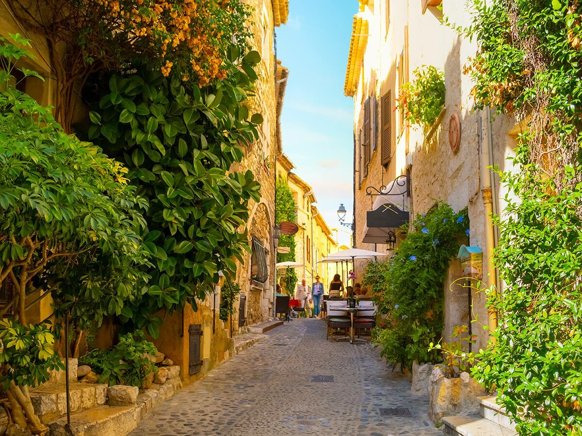 Malerische Gasse in südeuropäischer Altstadt mit historischen Gebäuden, vielen Pflanzen und Café-Tischen im Freien an einem sonnigen Tag.