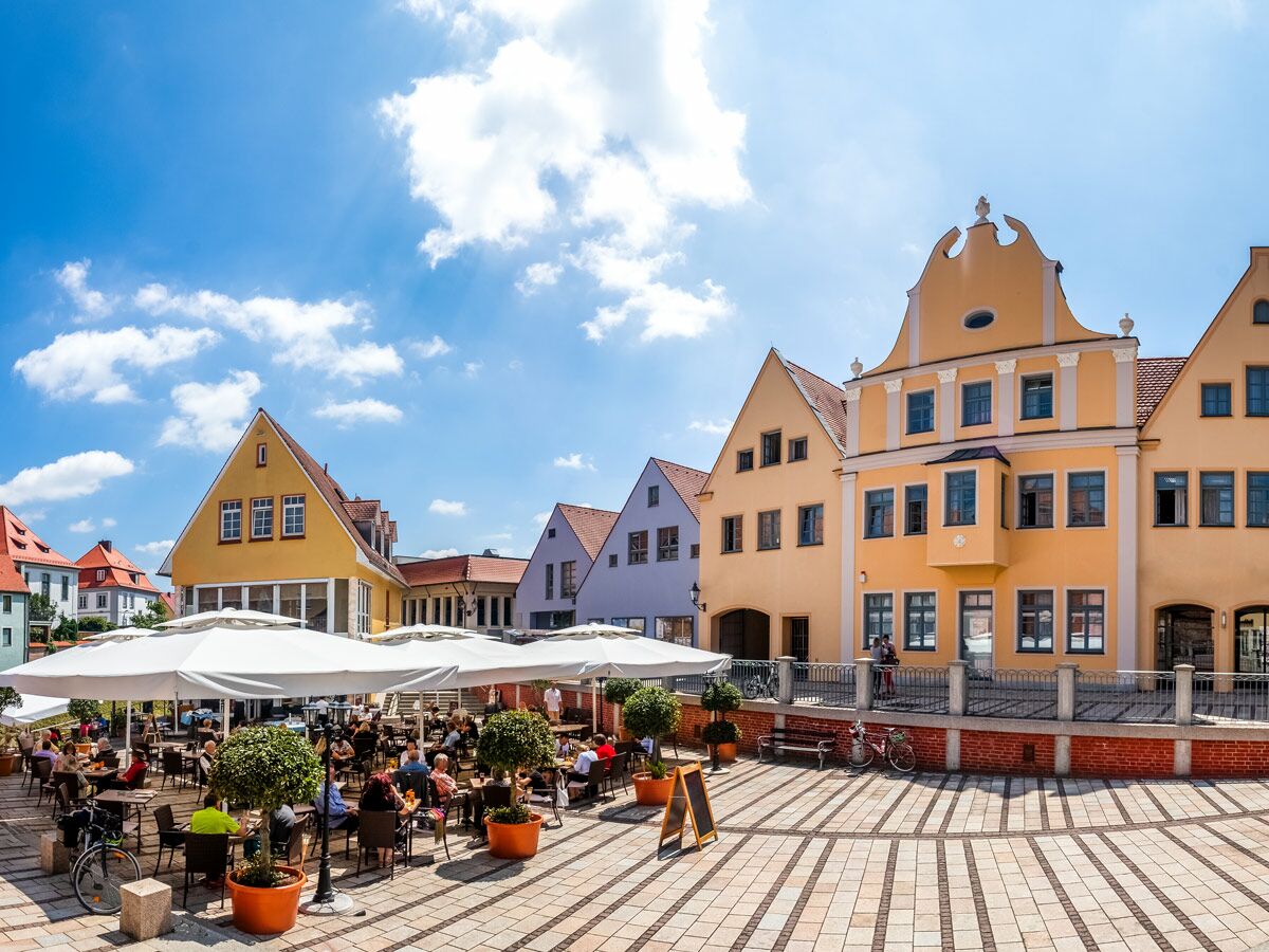 Belebtes Straßencafé auf einem sonnigen Stadtplatz mit farbigen historischen Giebelhäusern und vielen Menschen.