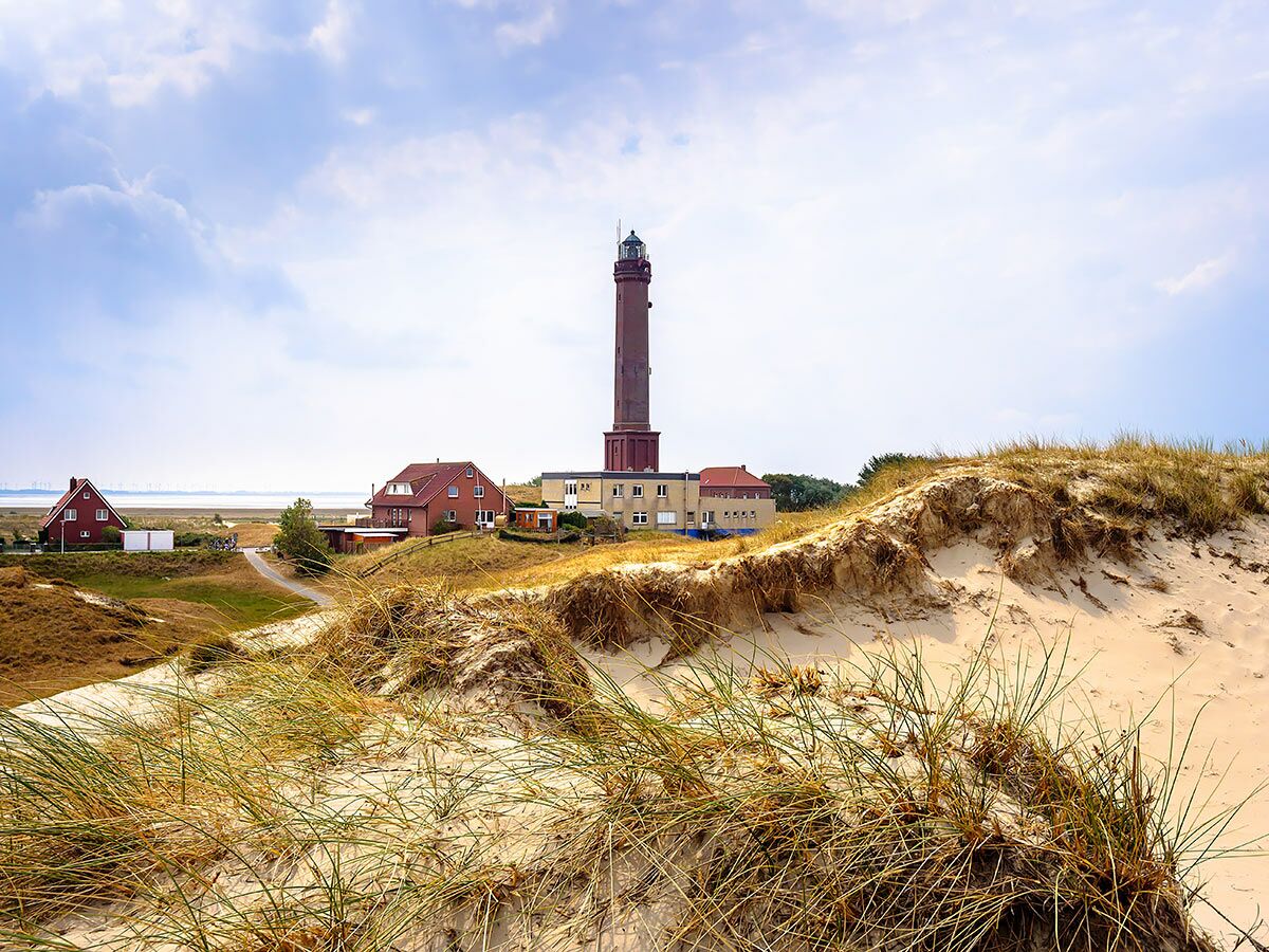 Roter Backstein-Leuchtturm mit Häusern in sandiger Dünenlandschaft an der Küste unter bewölktem Himmel, Windräder am Horizont.