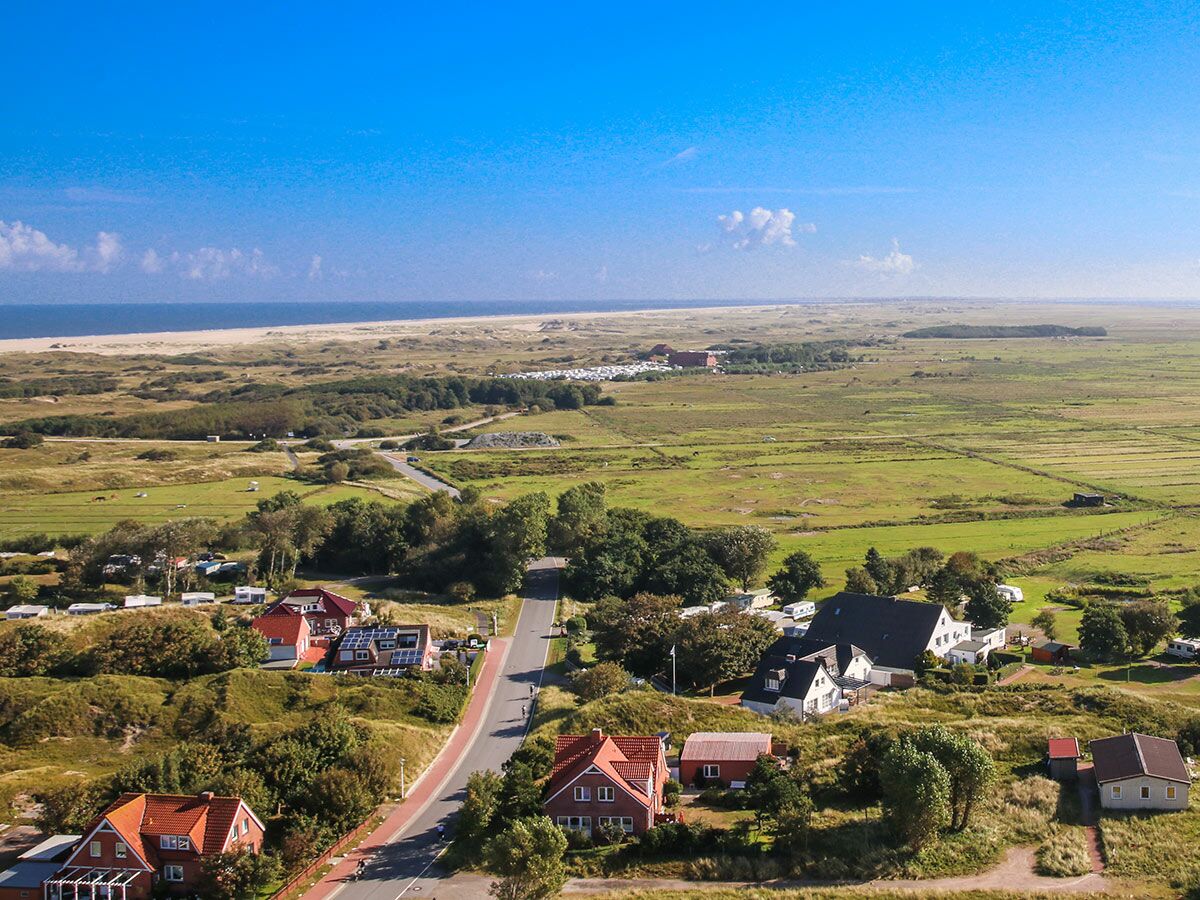 Luftaufnahme einer weiten Küstenlandschaft mit Strand, Meer, Dünen, grünen Feldern und Häusern eines Küstendorfes unter blauem Himmel.