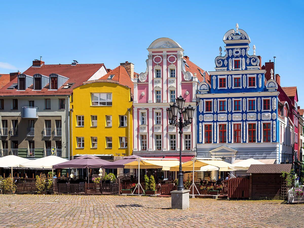 Reihe bunter Giebelhäuser an einem historischen Marktplatz mit Cafés und Kopfsteinpflaster unter blauem Himmel.