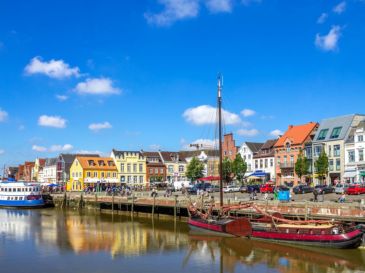 Ansicht einer europäischen Hafenstadt mit bunten Häusern entlang eines Kanals, historischen Schiffen und strahlend blauem Himmel.