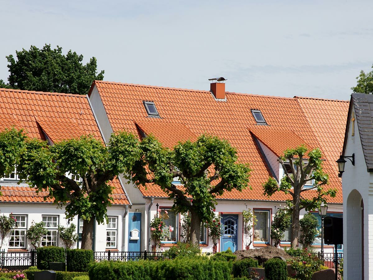 Reihe weißer Häuser mit orangen Ziegeldächern, blauen Türen, gepflegten Bäumen und Hecken unter blauem Himmel.