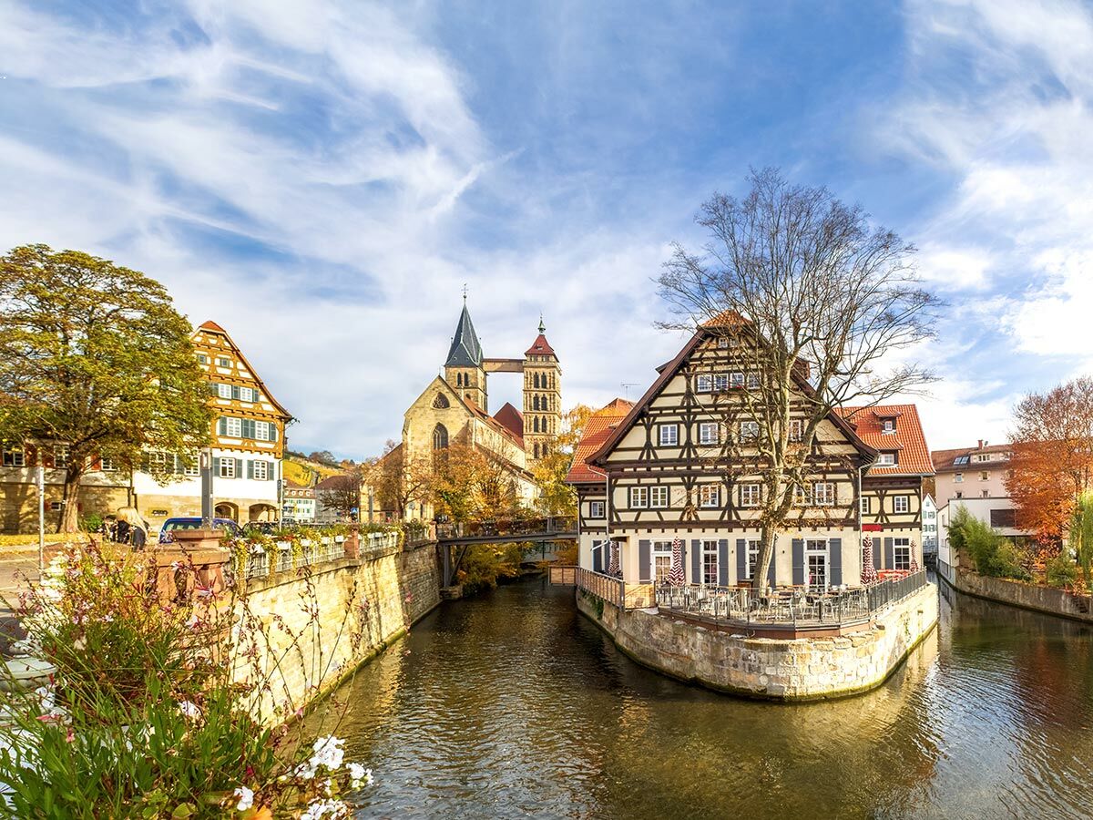 Malerische Neckarfront in Tübingen mit Fachwerkhäusern am Fluss, Stiftskirche und bunten Herbstbäumen unter blauem Himmel.