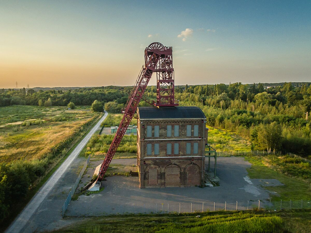 Historisches Zechengebäude mit rotem Förderturm im Abendlicht, umgeben von grünen Feldern und Bäumen, bei Sonnenuntergang.