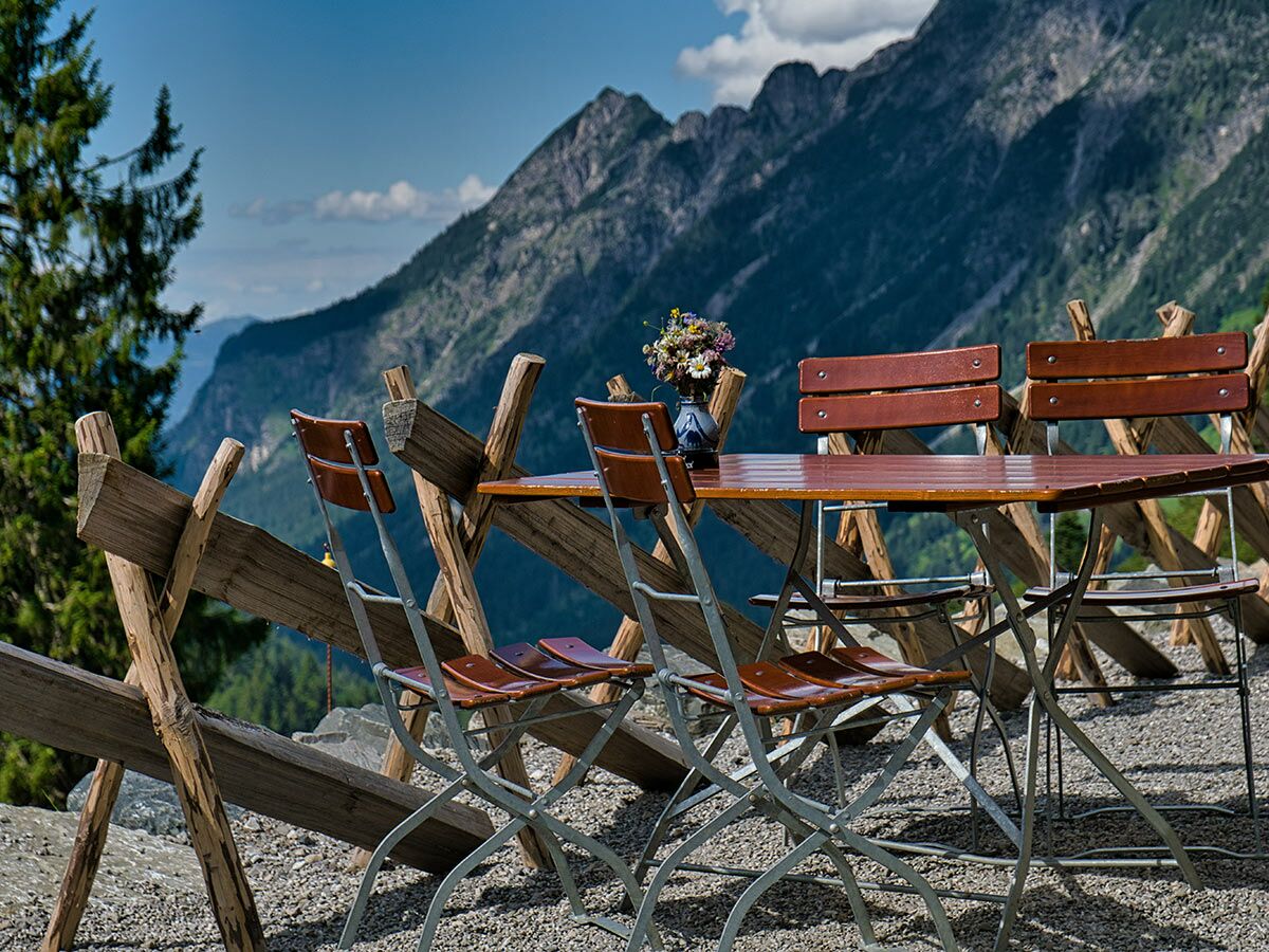 Sonniger Bergblick mit leeren Holztischen und Stühlen im Freien, bereit für Gäste, um die Alpenlandschaft zu genießen.