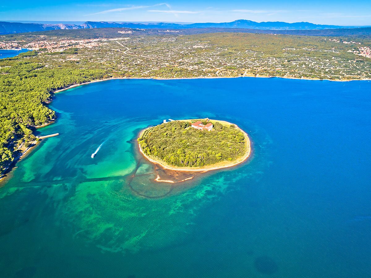 Luftaufnahme einer bewaldeten Insel mit alter Festung in einer türkisfarbenen Bucht, umgeben von klarem Meer und Küstenlandschaft.