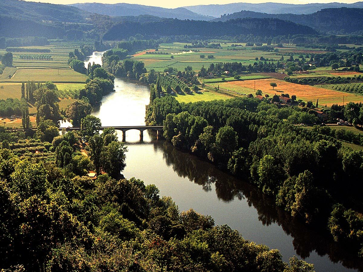 Weite Panoramablick auf Flusslandschaft mit Brücke, grünen Feldern und bewaldeten Hügeln unter klarem Himmel in Frankreich.