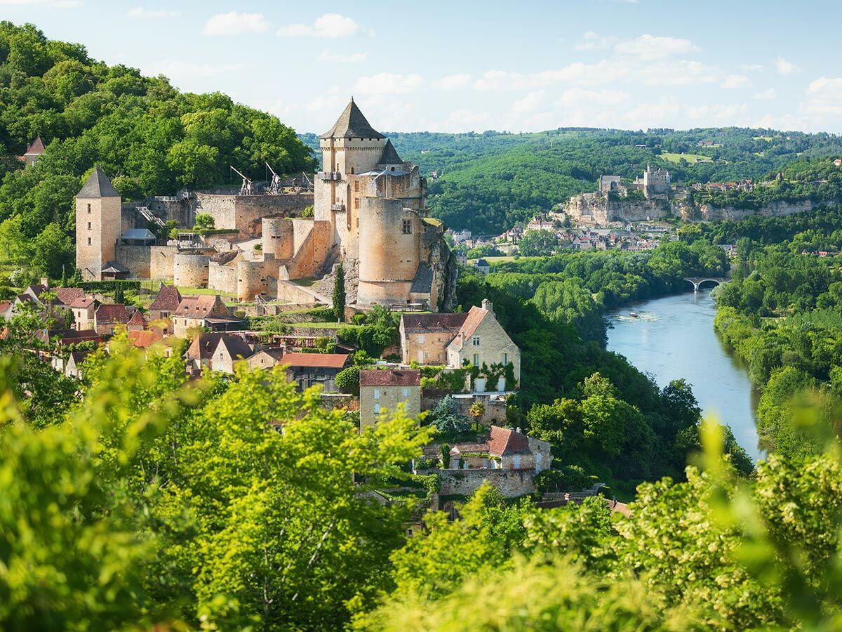 Panoramablick auf mittelalterliches Dorf und Burg am Fluss Dordogne, Frankreich, umgeben von grünen Hügeln.