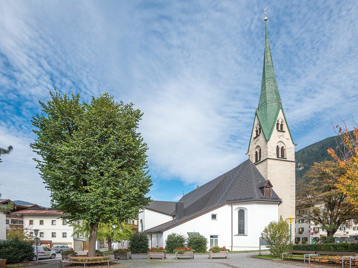 Helle Alpenkirche mit grünem Spitzturm, großem Baum und Bänken auf Dorfplatz vor Bergkulisse bei blauem Himmel.