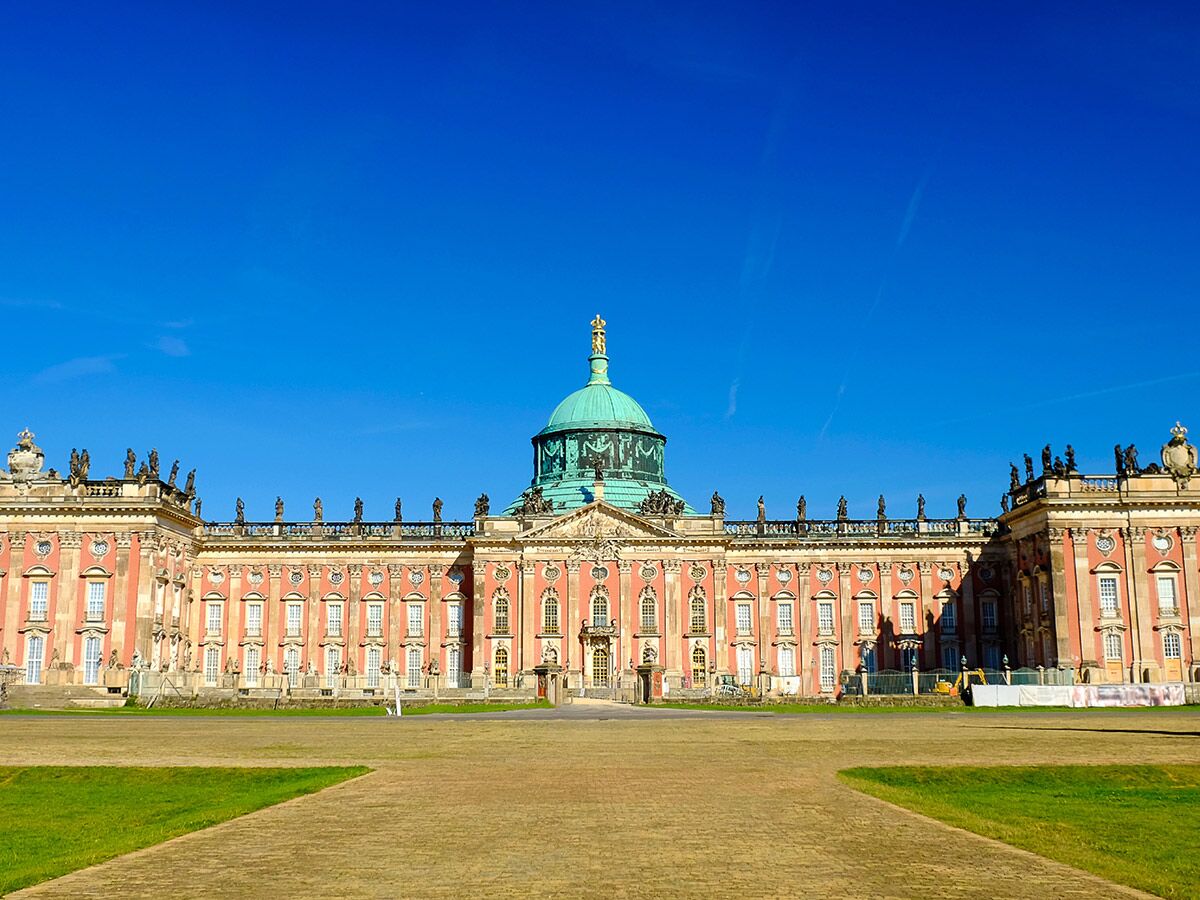 Barockes Neues Palais in Potsdam mit grüner Kuppel, Statuen und weitläufigem Rasen unter blauem Himmel.