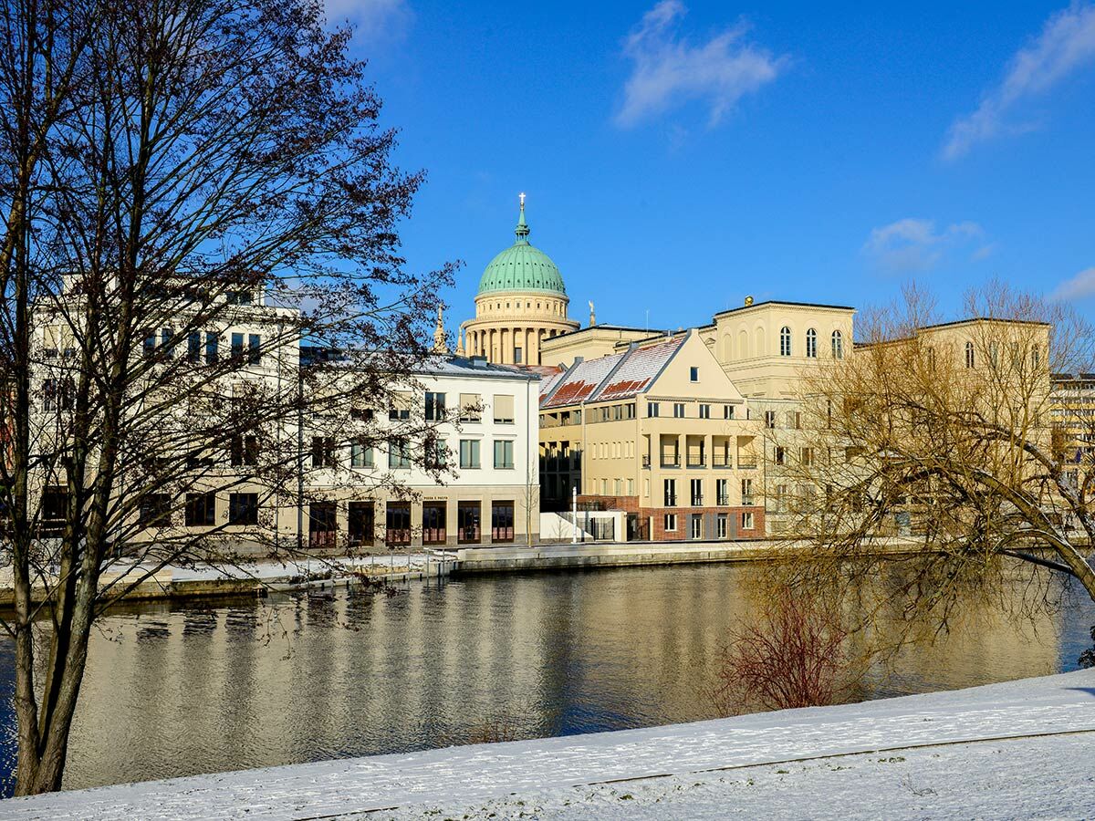 Winterliches Stadtbild von Potsdam mit Nikolaikirche, Havelufer und verschneitem Boden unter blauem Himmel.