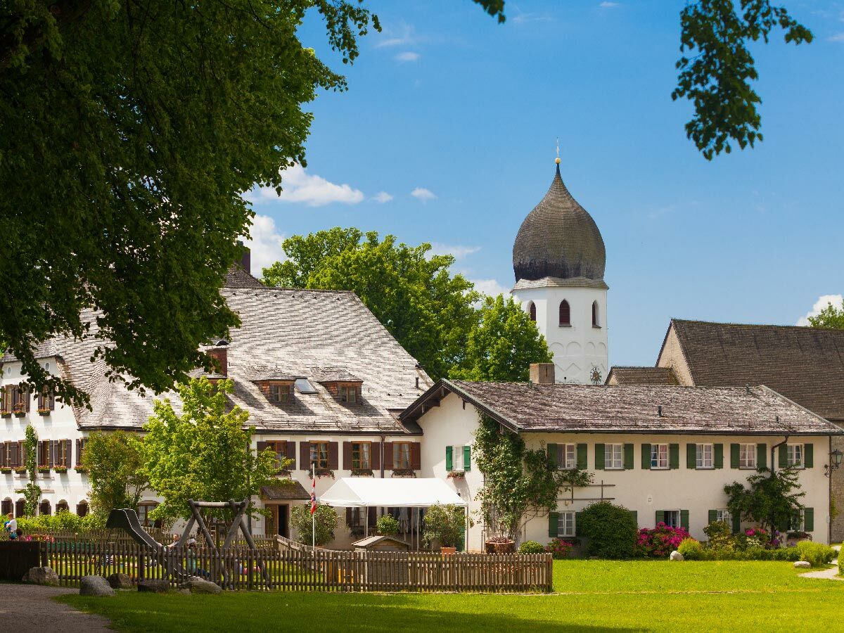 Idyllisches bayerisches Dorf mit traditionellen Häusern, Holzzaun, Spielplatz und markantem Zwiebelturm unter blauem Himmel.