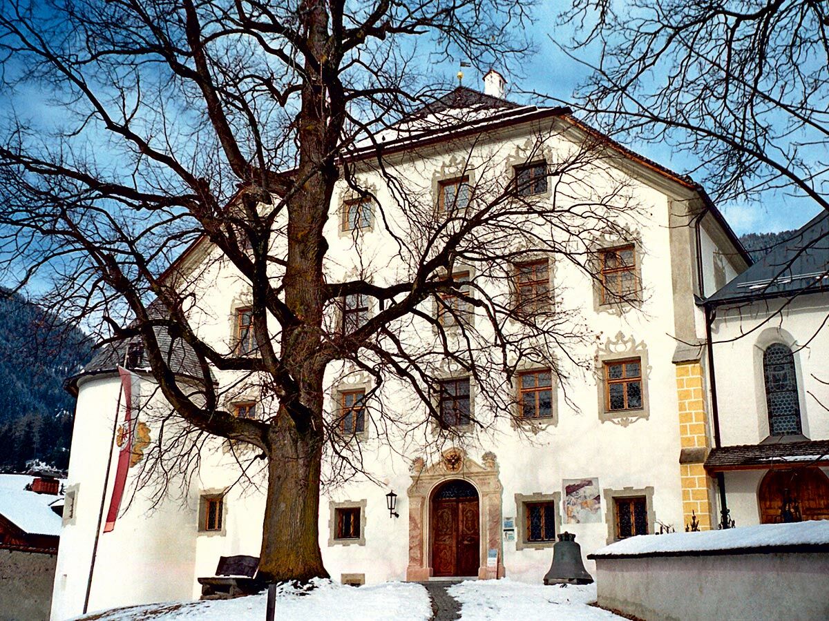 Weißes historisches Gebäude mit großem kahlen Baum davor, bedeckt mit Schnee, Berglandschaft unter blauem Himmel.