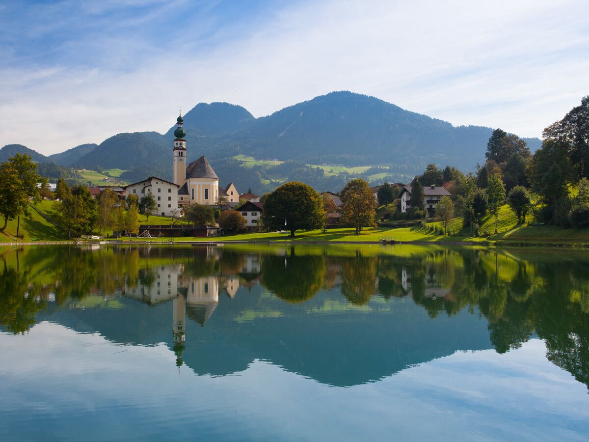 Malerisches Bergdorf mit Kirche und spiegelndem See, umgeben von grünen Hügeln und Bergen unter blauem Himmel.