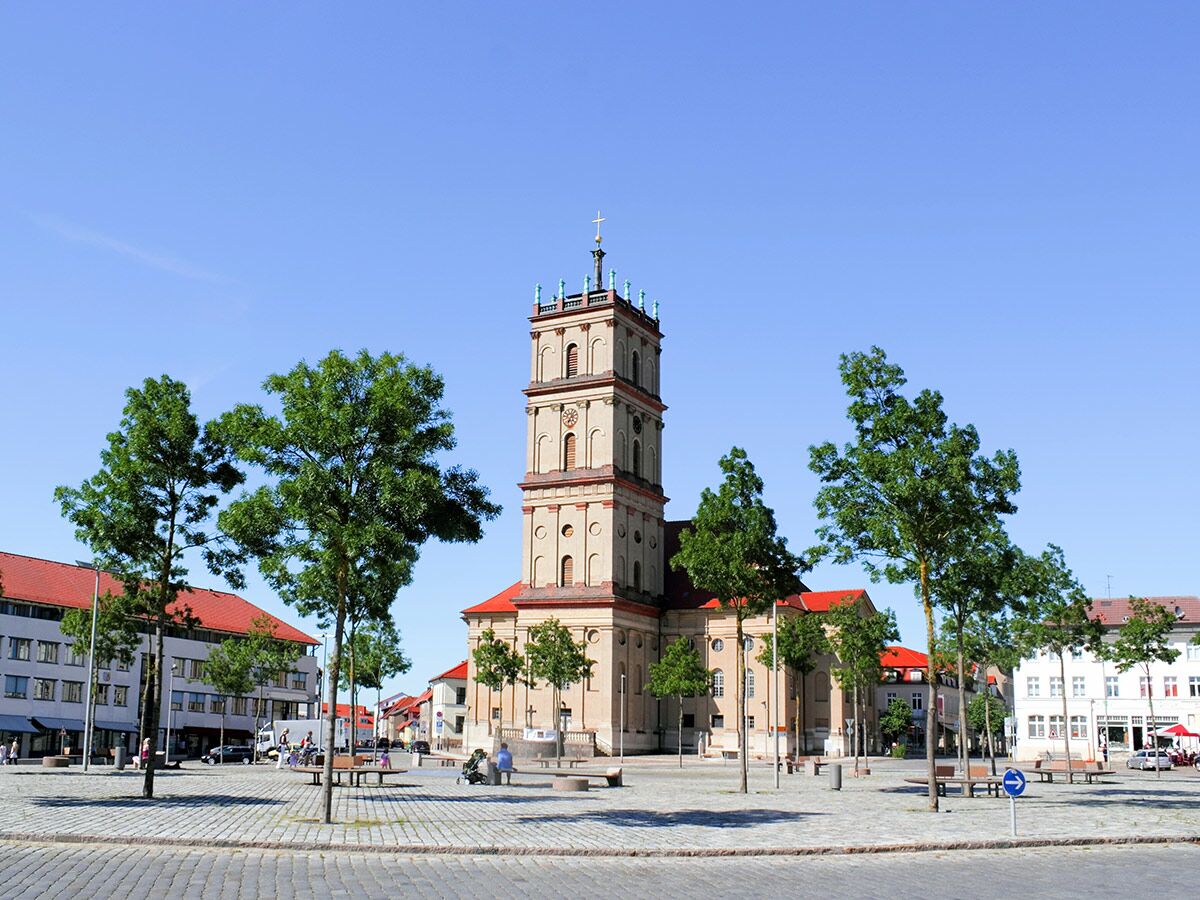 Großer historischer Kirchturm auf einem sonnigen, gepflasterten Marktplatz mit grünen Bäumen und Menschen.
