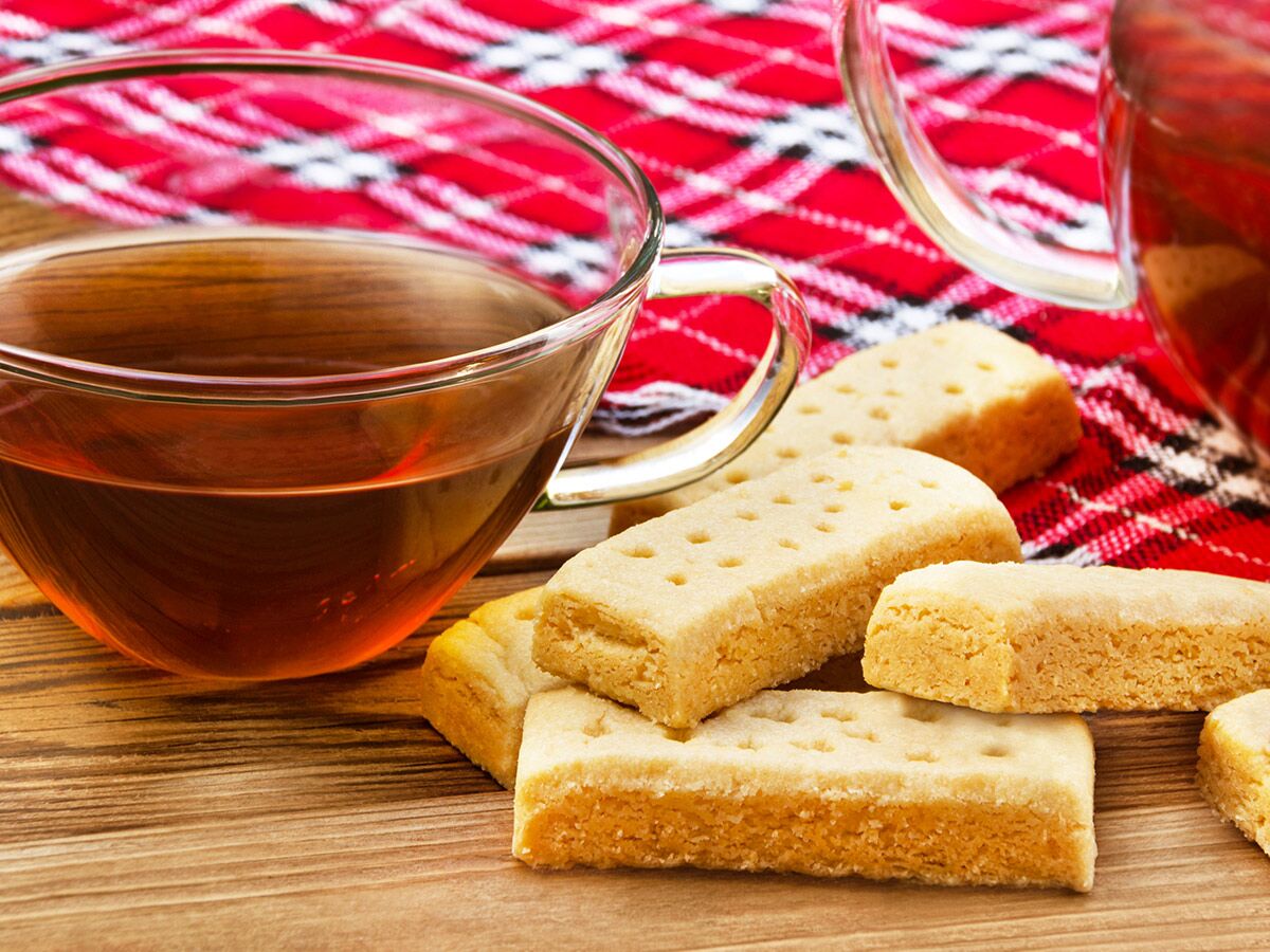 Gemütliche Teestunde mit einer Tasse Schwarztee, Teekanne und leckeren Shortbread-Keksen auf Holztisch und karierter Decke.