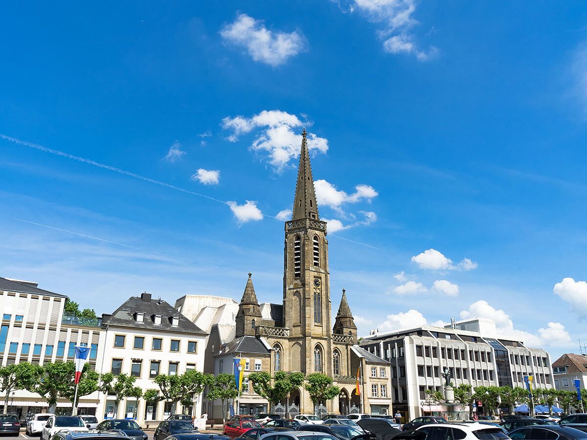 Hohe Kirche mit Turm auf einem sonnigen Stadtplatz, umgeben von Gebäuden und parkenden Autos unter blauem Himmel.
