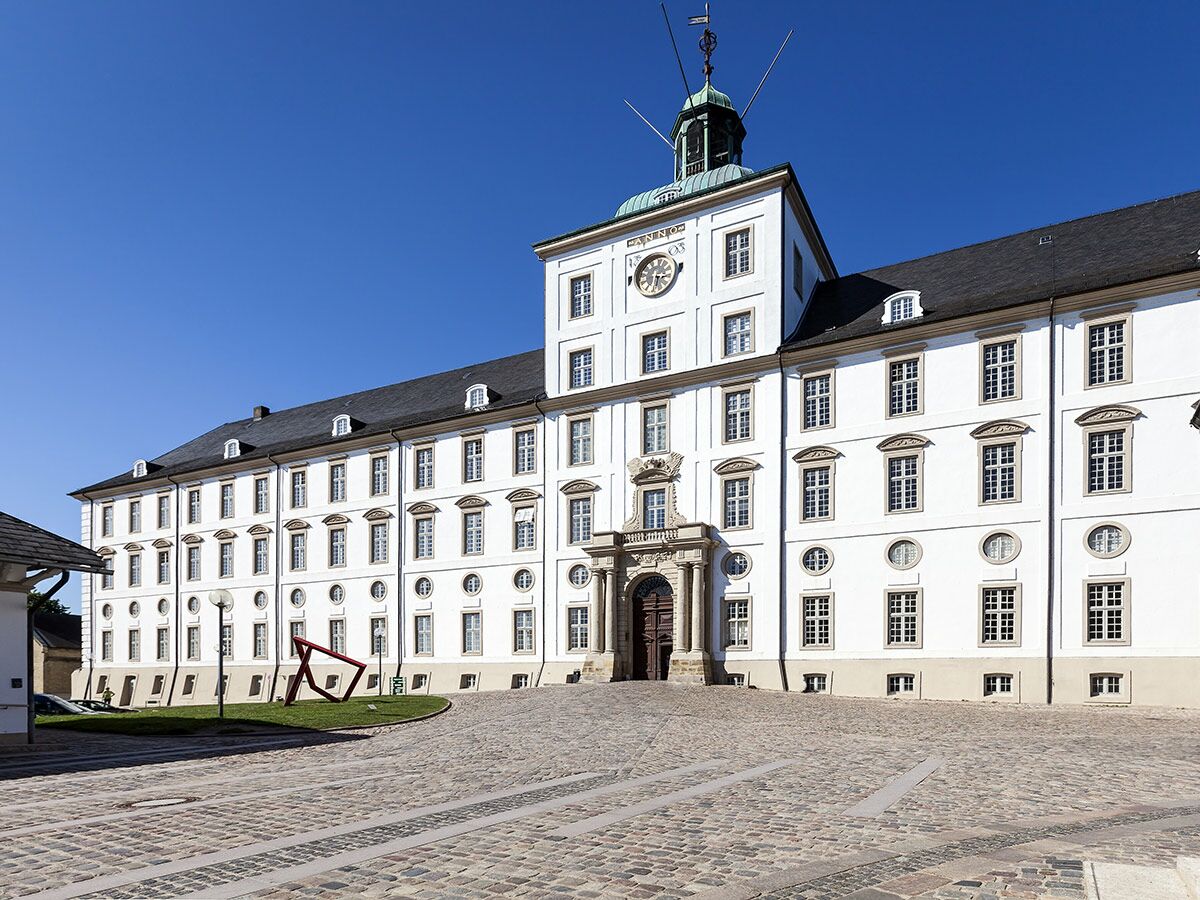 Historisches Schloss Corvey mit weißer Fassade, Uhrturm und grünem Dach, davor Kopfsteinpflaster und rote Skulptur unter blauem Himmel.