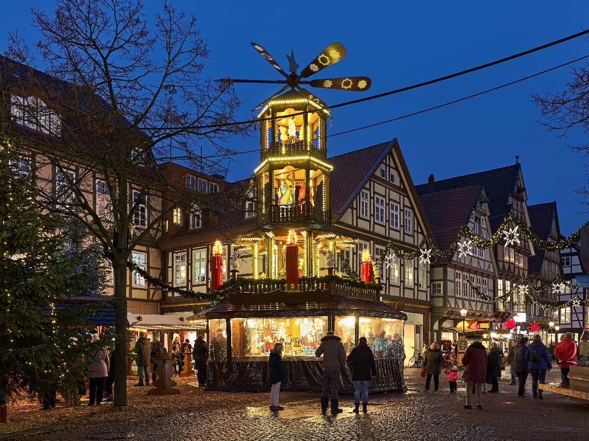 Beleuchteter Weihnachtsmarkt mit großer Weihnachtspyramide, Fachwerkhäusern und Menschen bei Dämmerung, festliche Atmosphäre.