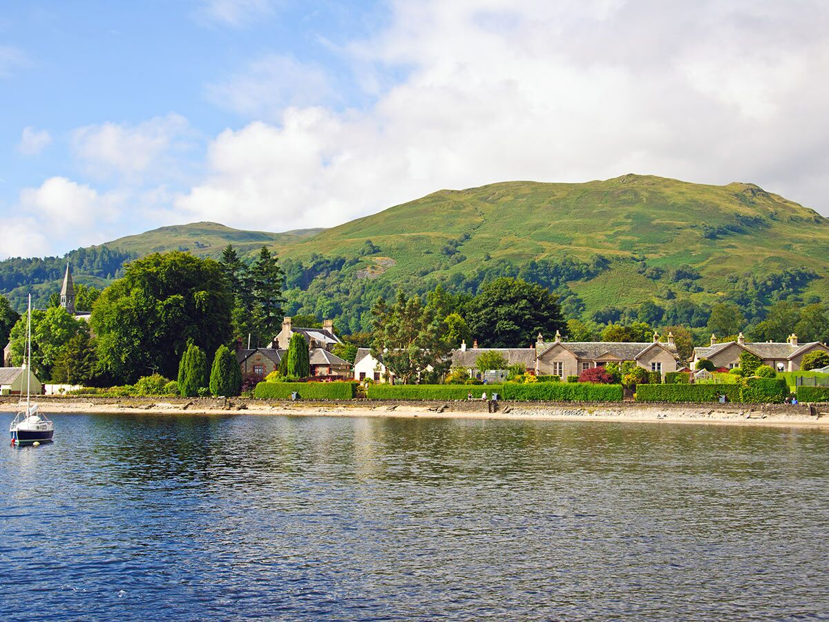 Idyllisches schottisches Dorf am Ufer eines blauen Sees mit grünen Bergen, Boot und Steinhäusern unter blauem Himmel.