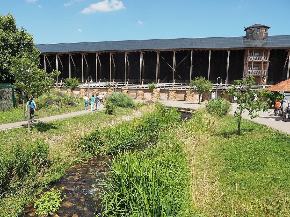 Besucher schlendern an einem sonnigen Tag entlang des langen Holz-Gradierwerks in einem Kurpark mit Fluss und Bäumen.