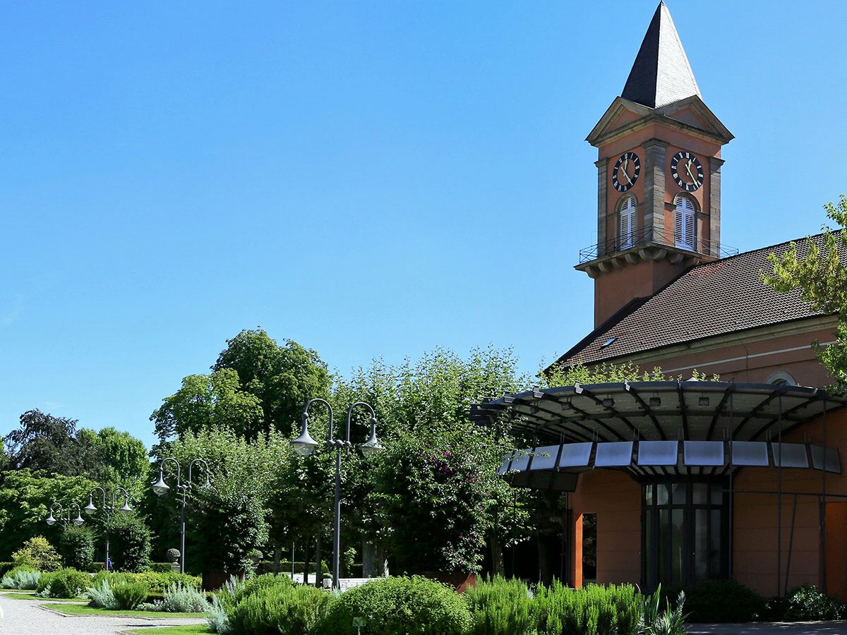 Historischer Kirchenturm mit Uhr, daneben modernes Eingangsgebäude, umgeben von einem grünen, sonnigen Park mit Laternen.