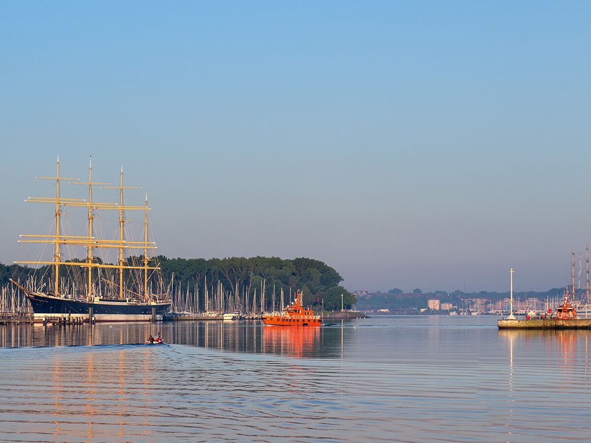 Großes Segelschiff im Hafen von Kiel, umgeben von Yachten, bei Sonnenaufgang mit einem kleinen Boot auf dem Wasser.