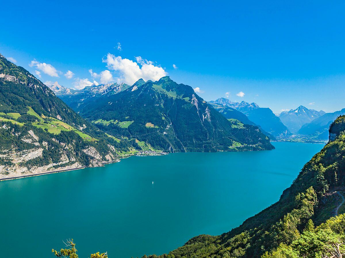 Türkisblauer Bergsee umgeben von grünen Alpenhängen, schneebedeckten Gipfeln und blauem Himmel mit Wolken, kleines Dorf.