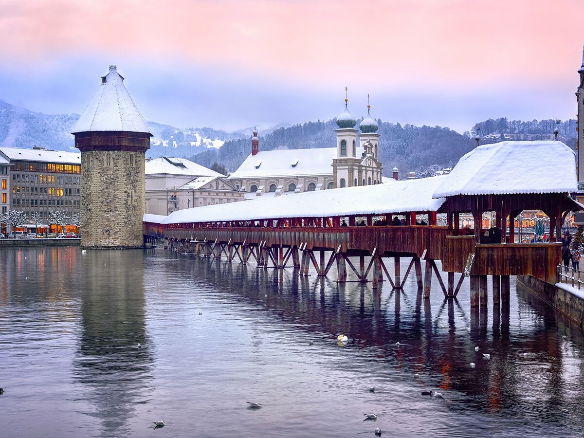 Historische Kapellbrücke und Wasserturm in Luzern, Schweiz, winterlich verschneit unter rosa Abendhimmel mit Bergen.