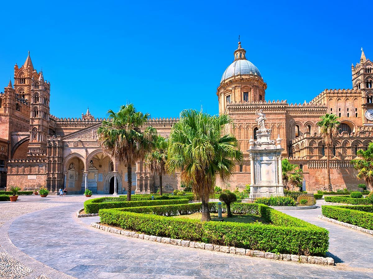 Palermo Kathedrale in Sizilien mit ihrer beeindruckenden Architektur, großer Kuppel, Türmen, Palmen und einem gepflegten Garten unter blauem Himmel.