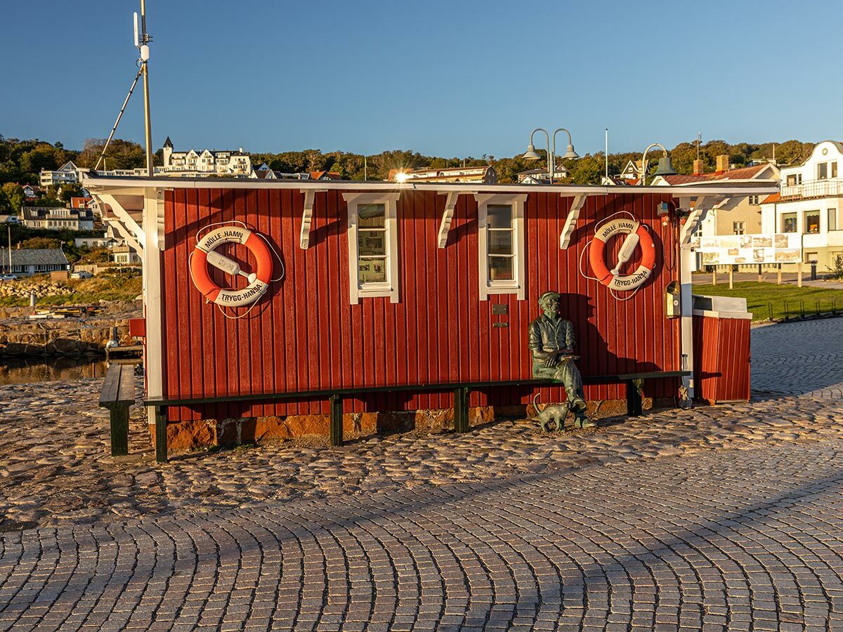 Rotes Holzgebäude mit sitzender Statue und Hund auf Bank, davor Pflastersteine, im Hintergrund Mölle Hafen und Häuser.