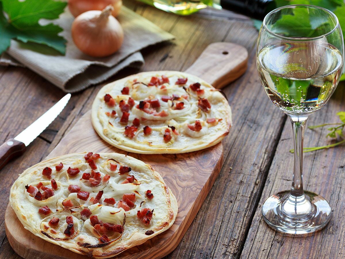 Zwei Flammkuchen mit Zwiebeln und Speck auf Holzbrett, daneben Weißwein und Zwiebeln.