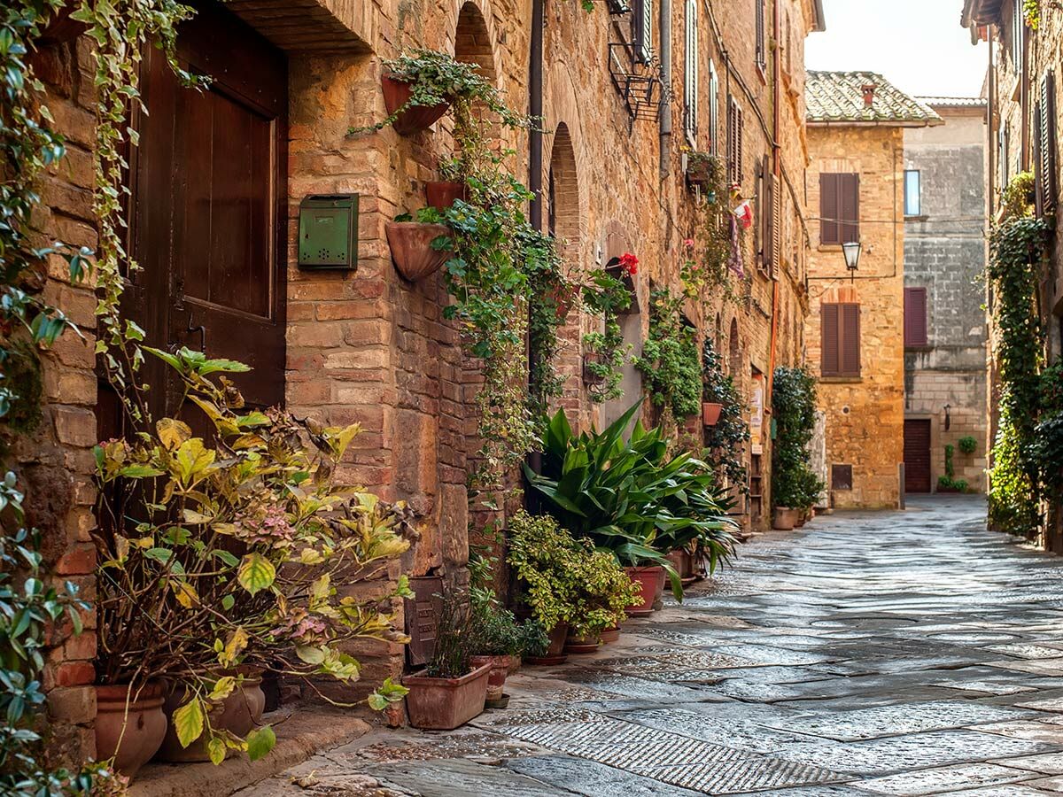 Enge Gasse in einer historischen italienischen Stadt mit Backsteinhäusern, Holztür, Blumentöpfen und rankenden Pflanzen.