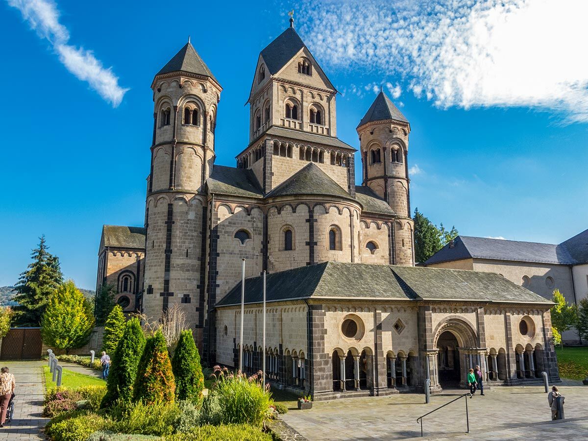 Majestätische romanische Abtei Maria Laach mit mehreren Türmen, in der Sonne leuchtend, umgeben von gepflegten Gärten.
