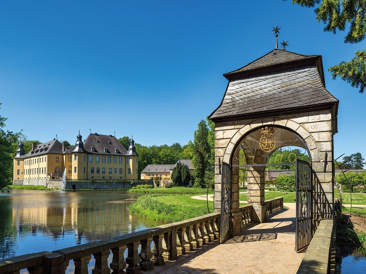 Historisches gelbes Wasserschloss mit Wassergraben, Springbrunnen, einer Steinbrücke und verziertem Tor in grüner Parkanlage.