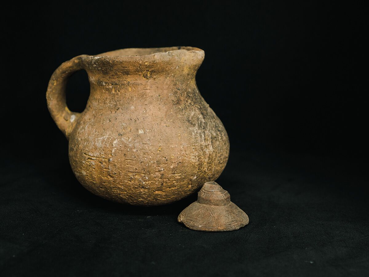 Alter, rauer Tonkrug mit Henkel und ein kleines konisches Tonartefakt auf schwarzem Hintergrund, archäologisches Fundstück.