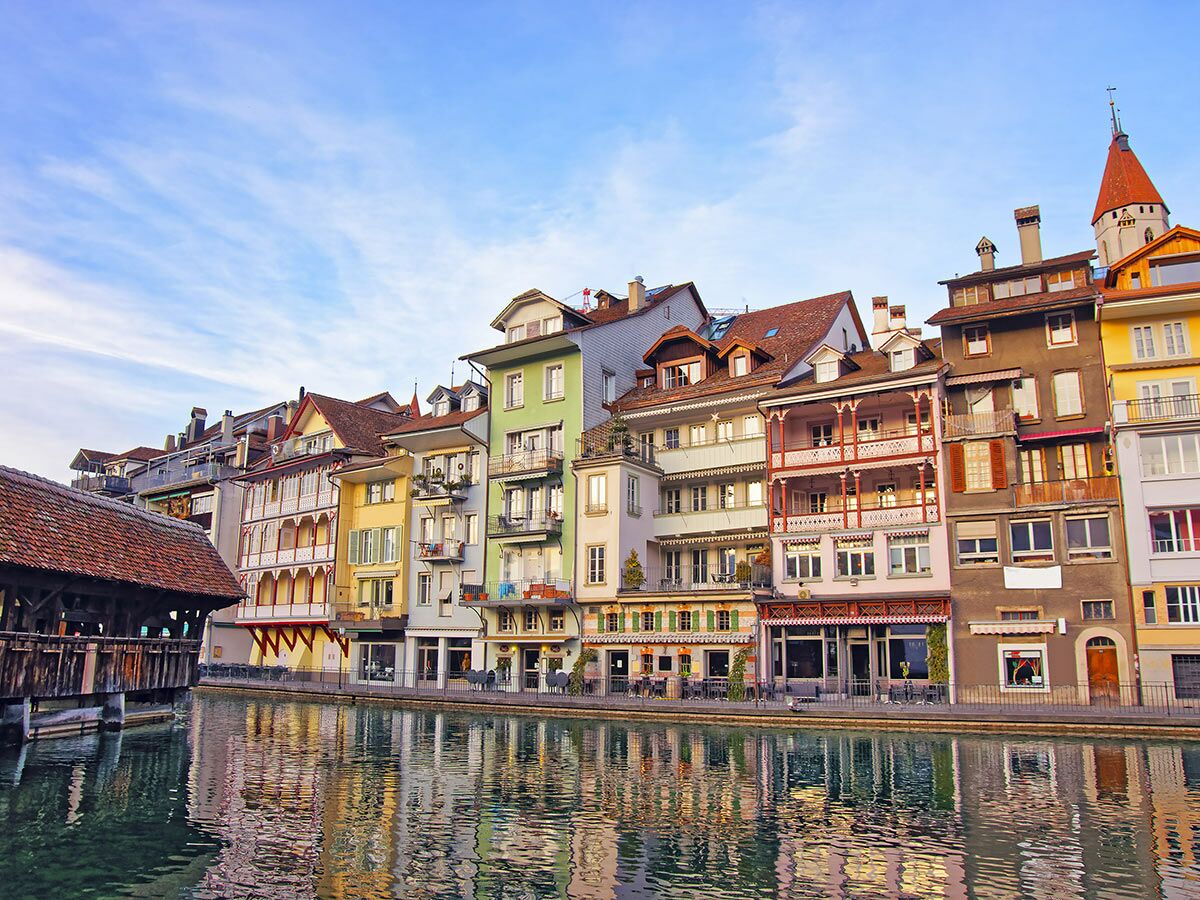 Bunte historische Häuserreihe am Fluss in Luzern mit überdachter Holzbrücke und Turm, gespiegelt im Wasser.
