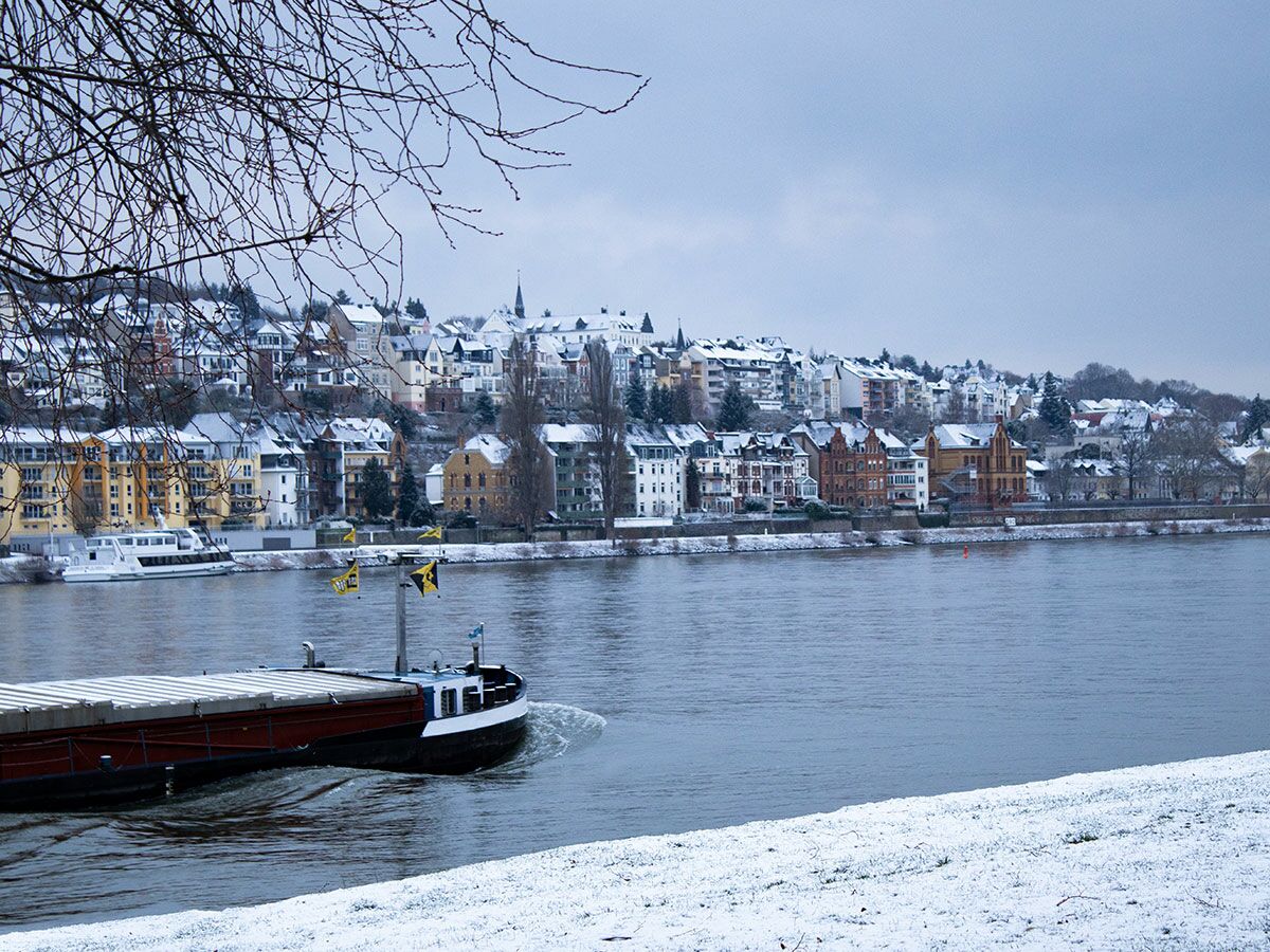 Schneebedeckte Stadt am Fluss mit Frachtschiff im Vordergrund an einem winterlichen Tag.