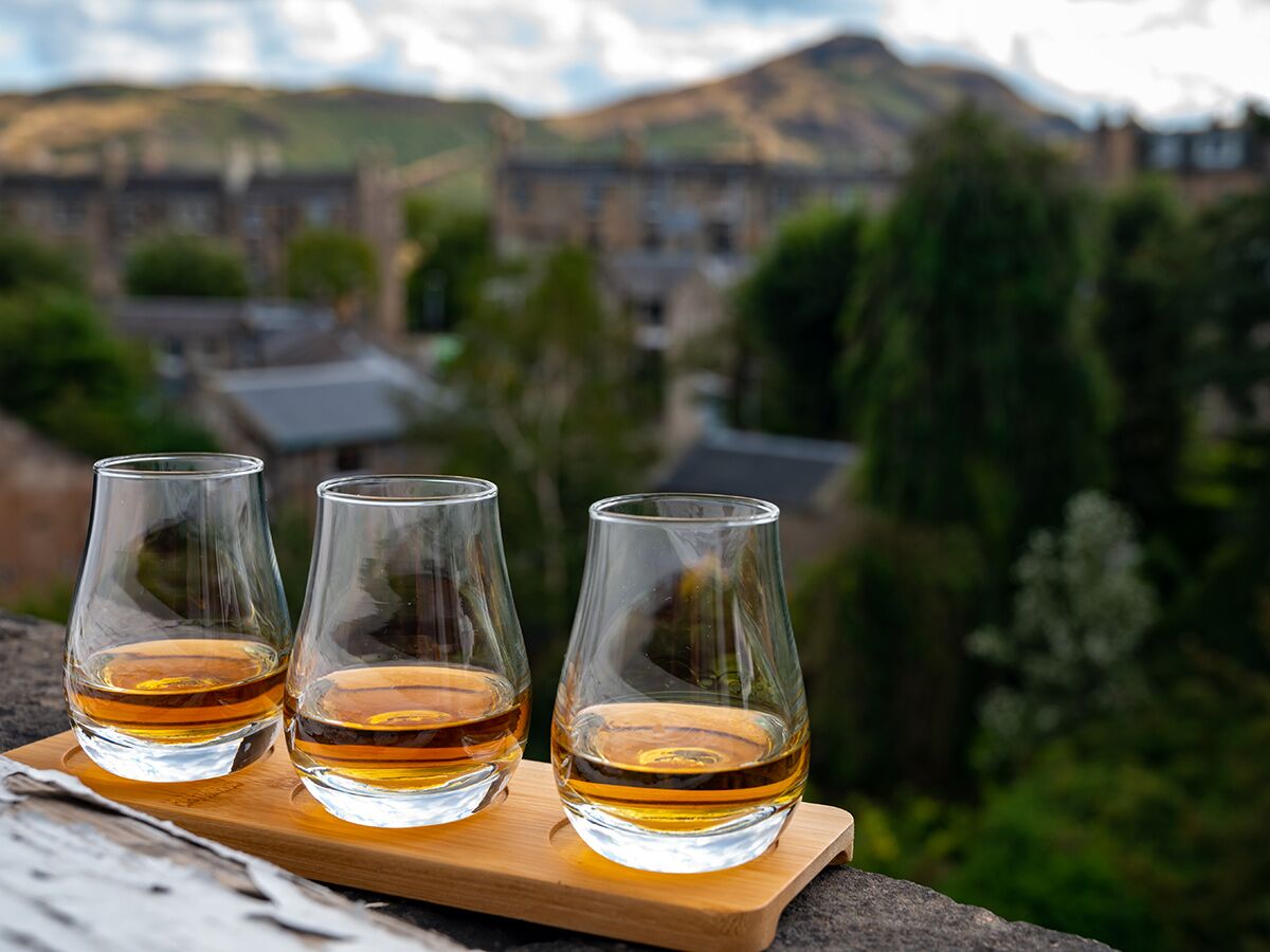Drei Gläser schottischer Whisky auf Holztablett mit verschwommenem Stadtblick von Edinburgh und Arthur's Seat.