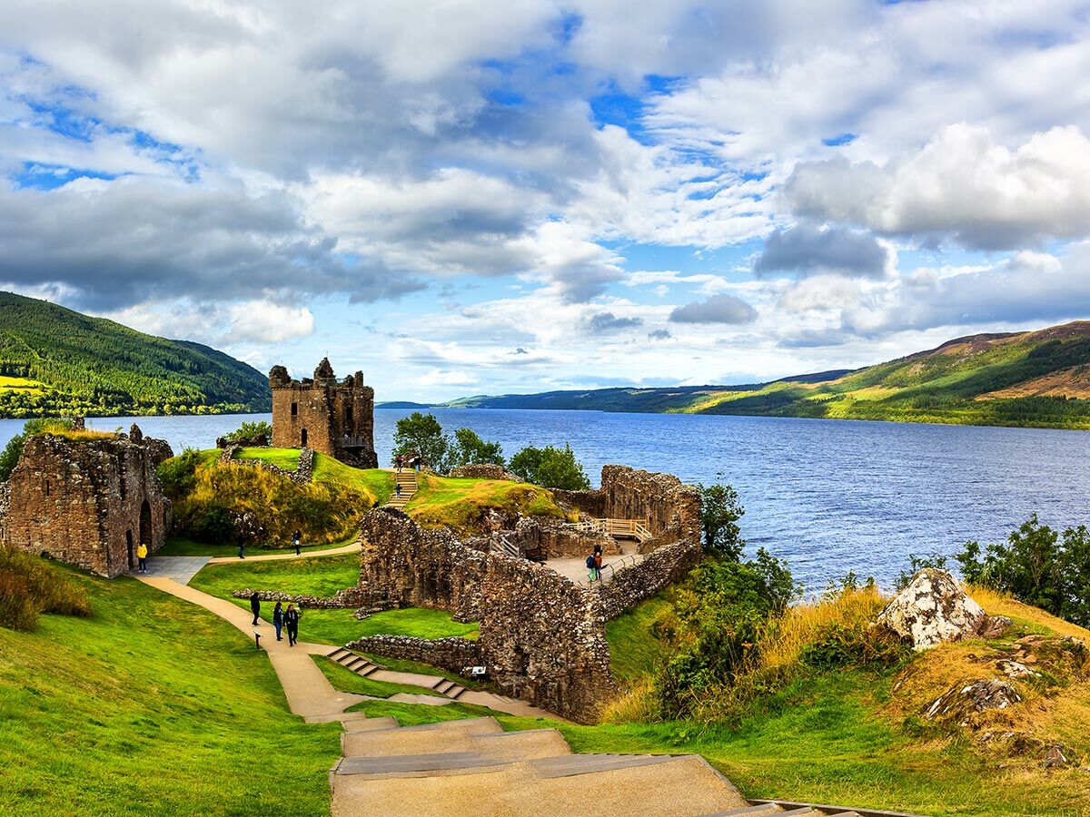 Panoramablick auf die historische Ruine Urquhart Castle am Ufer des Loch Ness mit grünen Hügeln und Wolkenhimmel.