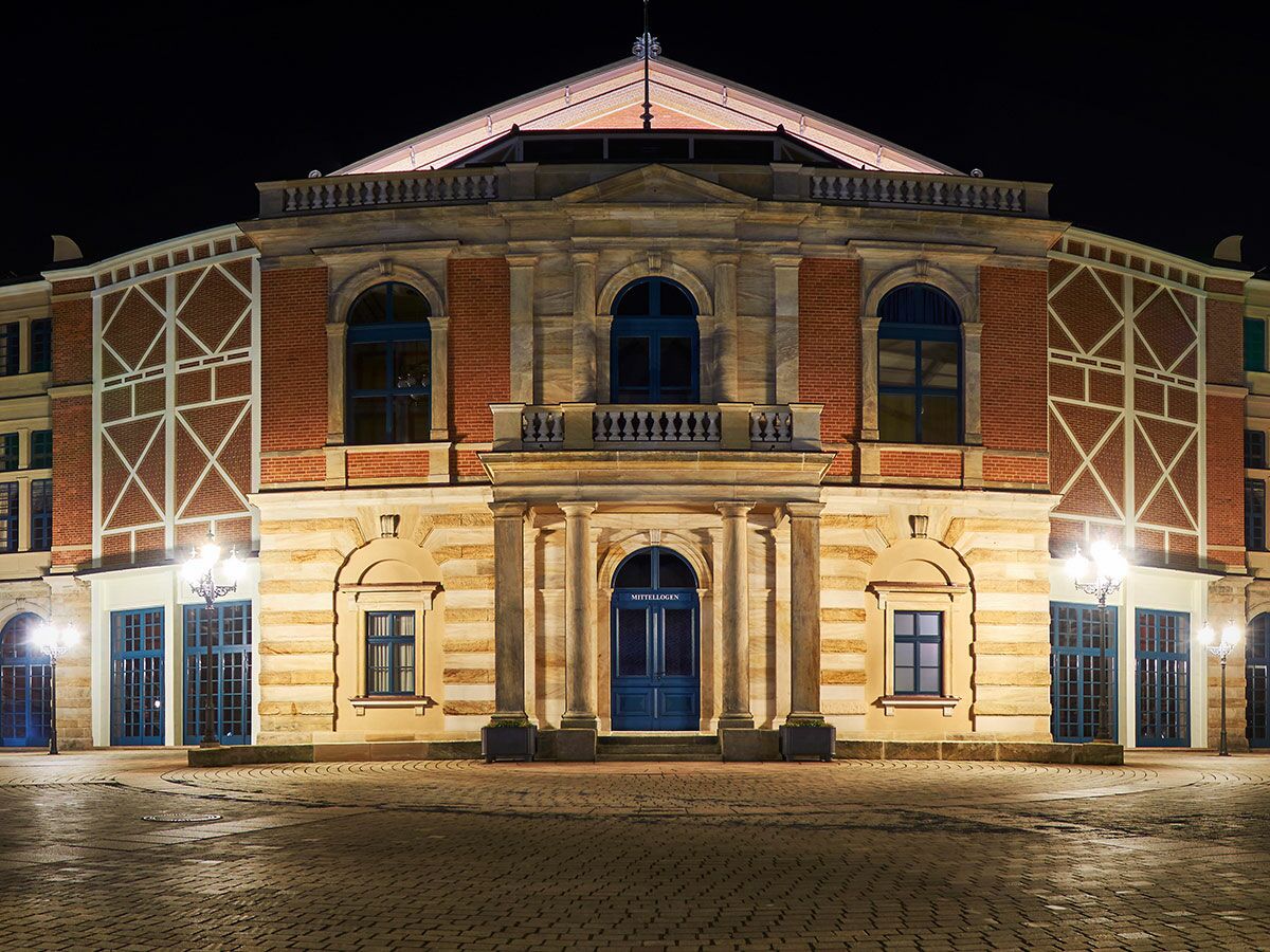 Nachtansicht des Bayreuther Festspielhauses, beleuchtet, mit klassischer Architektur, roten Ziegeln und blauen Türen. Kopfsteinpflaster im Vordergrund.