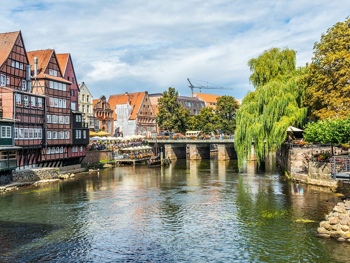 Lüneburgs historische Altstadt mit Fachwerkhäusern, Cafés am Flussufer, einer Brücke und einer malerischen Trauerweide an einem sonnigen Tag.